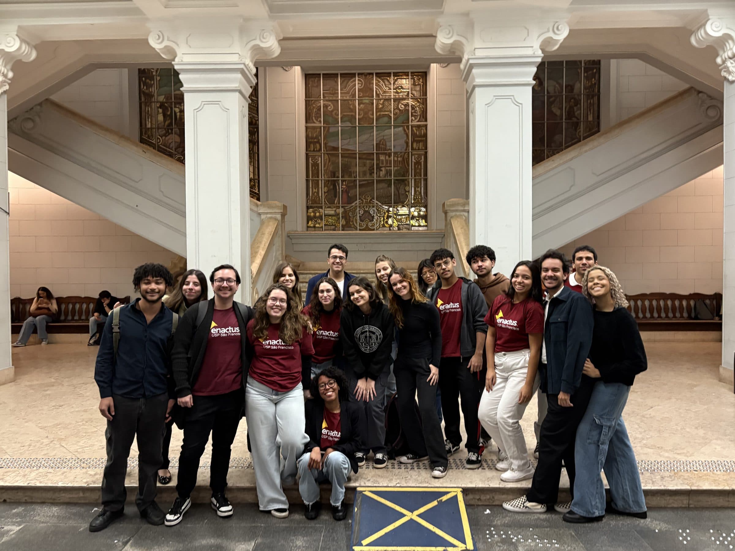 Membros da Enactus USP São Francisco, com as camisetas da equipe, reunidos no saguão da Faculdade de Direito da USP, em frente ao vitral